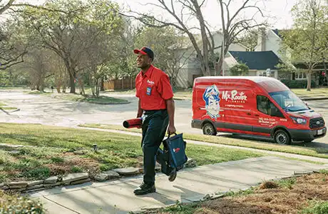 A Mr. Rooter plumber walking up path to a client's house with branded van parked on the street behind him.