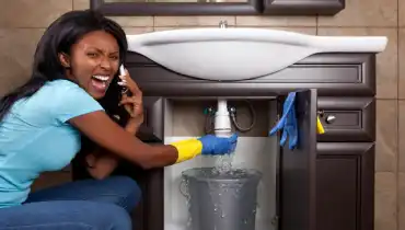A panicked woman is on the phone calling for plumbing assistance as a pipe below her sink gushes water.