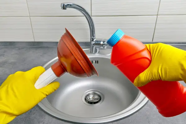 POV of a clogged sink drain with two hands, one holding a plunger and the other a bottle of drain cleaner