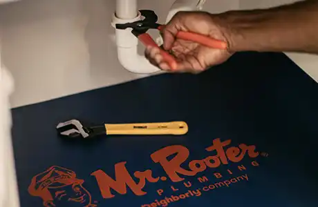 Mr. Rooter plumber's hand using a wrench to fix a pipe, with a Mr. Rooter mat underneath the pipes.