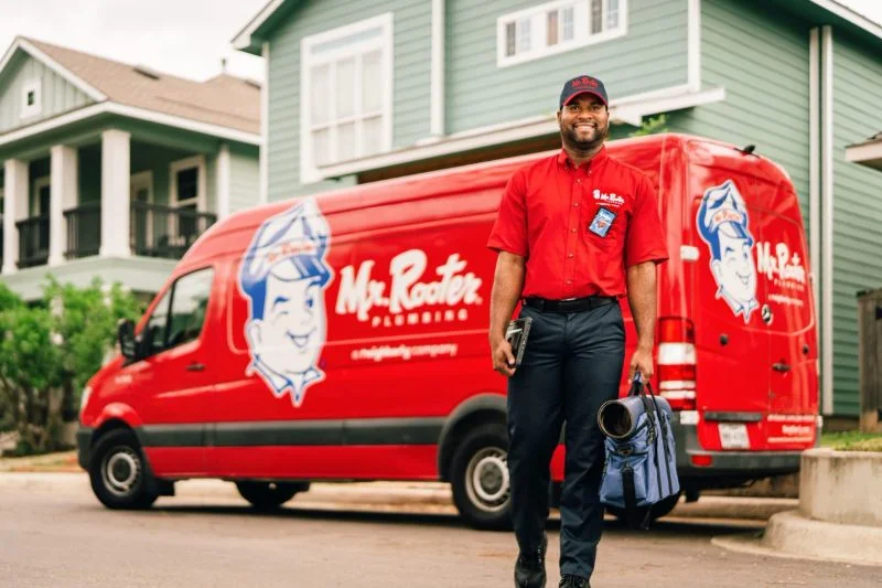 Mr. Rooter plumber standing in front of branded company van and two homes, smiling and holding work bag.