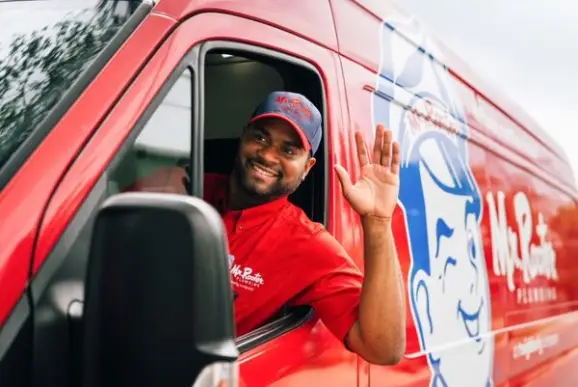 Mr. Rooter service professional driving branded company van and waving out the van window while smiling.