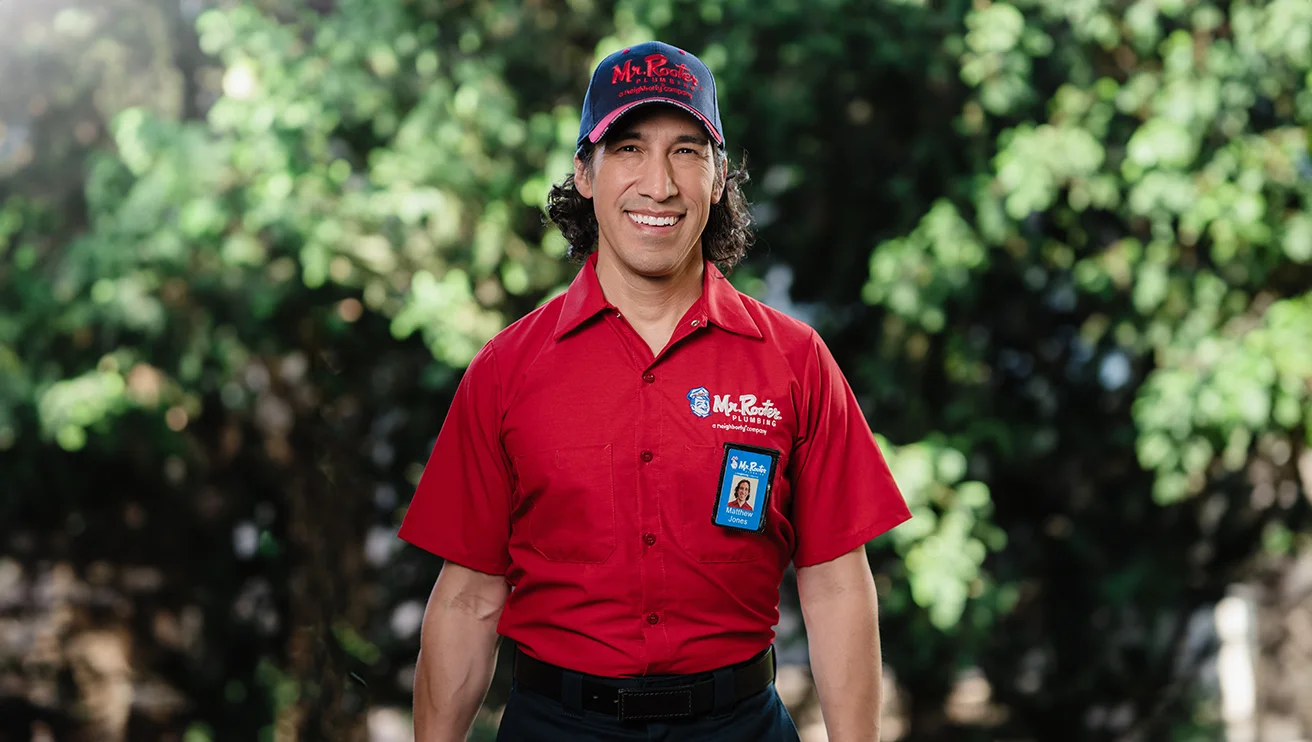 Smiling Mr. Rooter technician standing in front of a background of greenery.