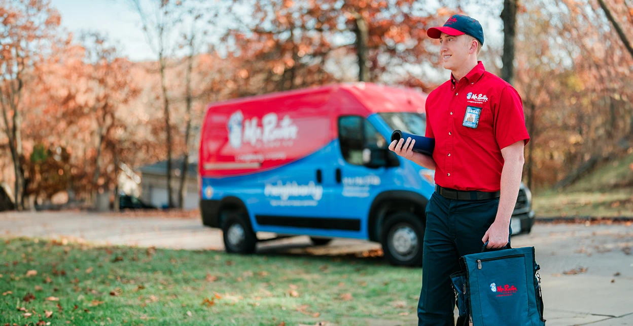 Mr. Rooter technician walking up to a customer's home with a branded work van parked in the background.