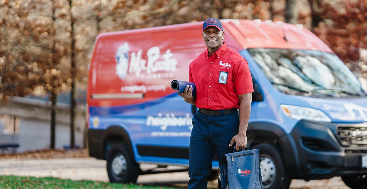 Mr. Rooter technician carrying a tool bag and standing beside a branded van.