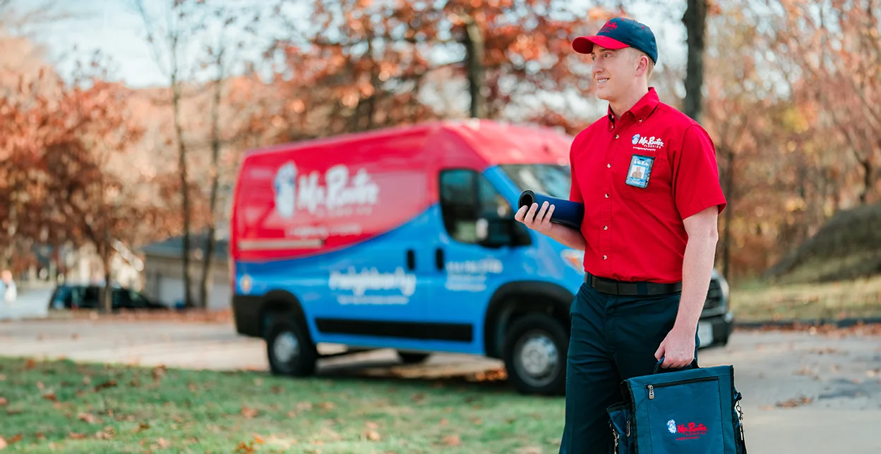 Mr. Rooter technician walking up to a customer's home with a branded van parked in the background.