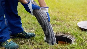 A plumbing technician guides a large plastic pipe into a hole in the ground where a septic tank is buried during the pumping process.