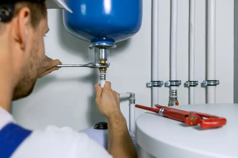 Professional plumber using a wrench to tighten fittings on a water heater expansion tank with visible pipes and plumbing tools.